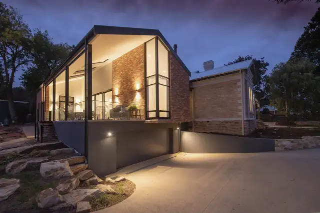 Small modern house at dusk with tall lit windows, a stone wall and a sloped garden