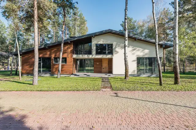 Low modern house with a pitched roof and cream panels among tall pines, a brick path leading to the door